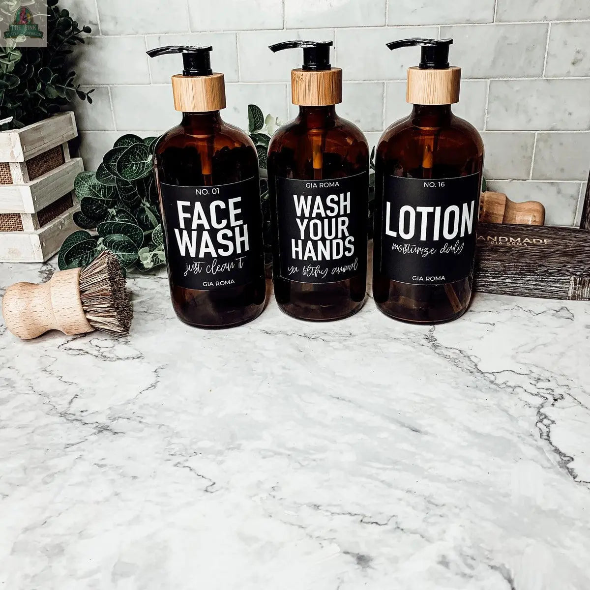 Three 16oz Glass Amber | Black Label bottles labeled “Face Wash,” “Wash Your Hands,” and “Lotion” sit on a marble countertop, accented by a wooden brush, greenery, and wood decor in the background.