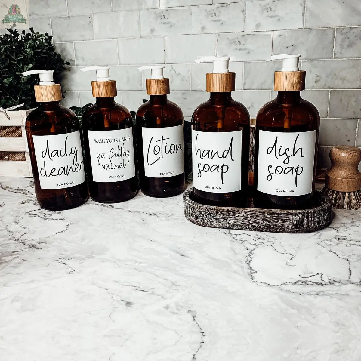 Five 16oz Glass Amber | White Label bottles labeled for daily cleaner, hand soap, dish soap, lotion, and a funny hand wash sit on a marble kitchen countertop with a tiled backsplash and wooden accents.