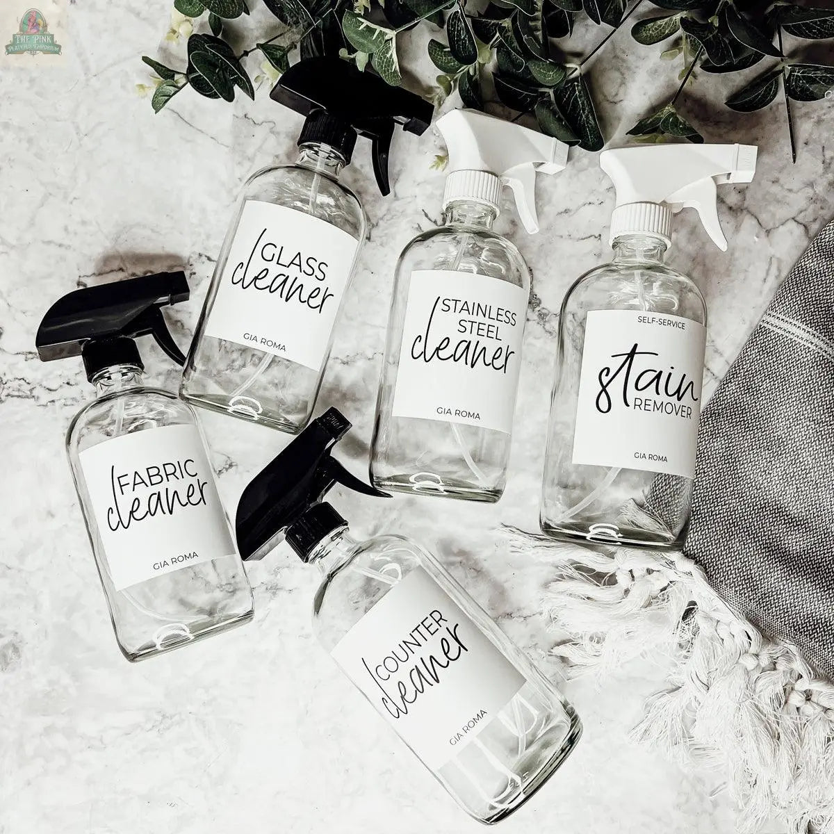 Five 16oz Glass Cleaning Bottles—Glass Cleaner, Stainless Steel Cleaner, Stain Remover, Fabric Cleaner, and Counter Cleaner—are arranged on a marble surface with greenery and a gray towel nearby.