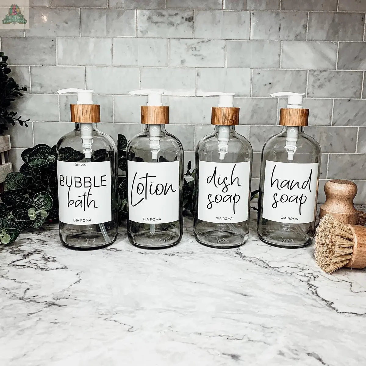 Four 16oz Glass Clear bottles with white labels and bamboo lids sit on a marble countertop, labeled “bubble bath,” “lotion,” “dish soap,” and “hand soap,” with nearby plants and wooden brushes.