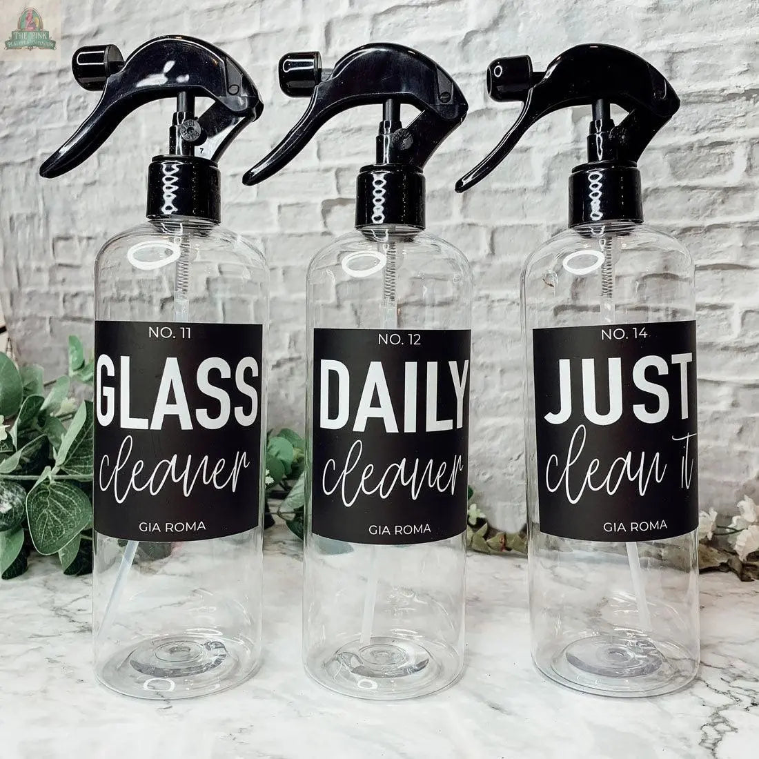 Three 16oz Plastic Cleaning Bottles with black spray tops, labeled “Glass Cleaner,” “Daily,” and “Just Clean It” with waterproof labels, sit on a white surface near a green plant and a light brick wall backdrop.