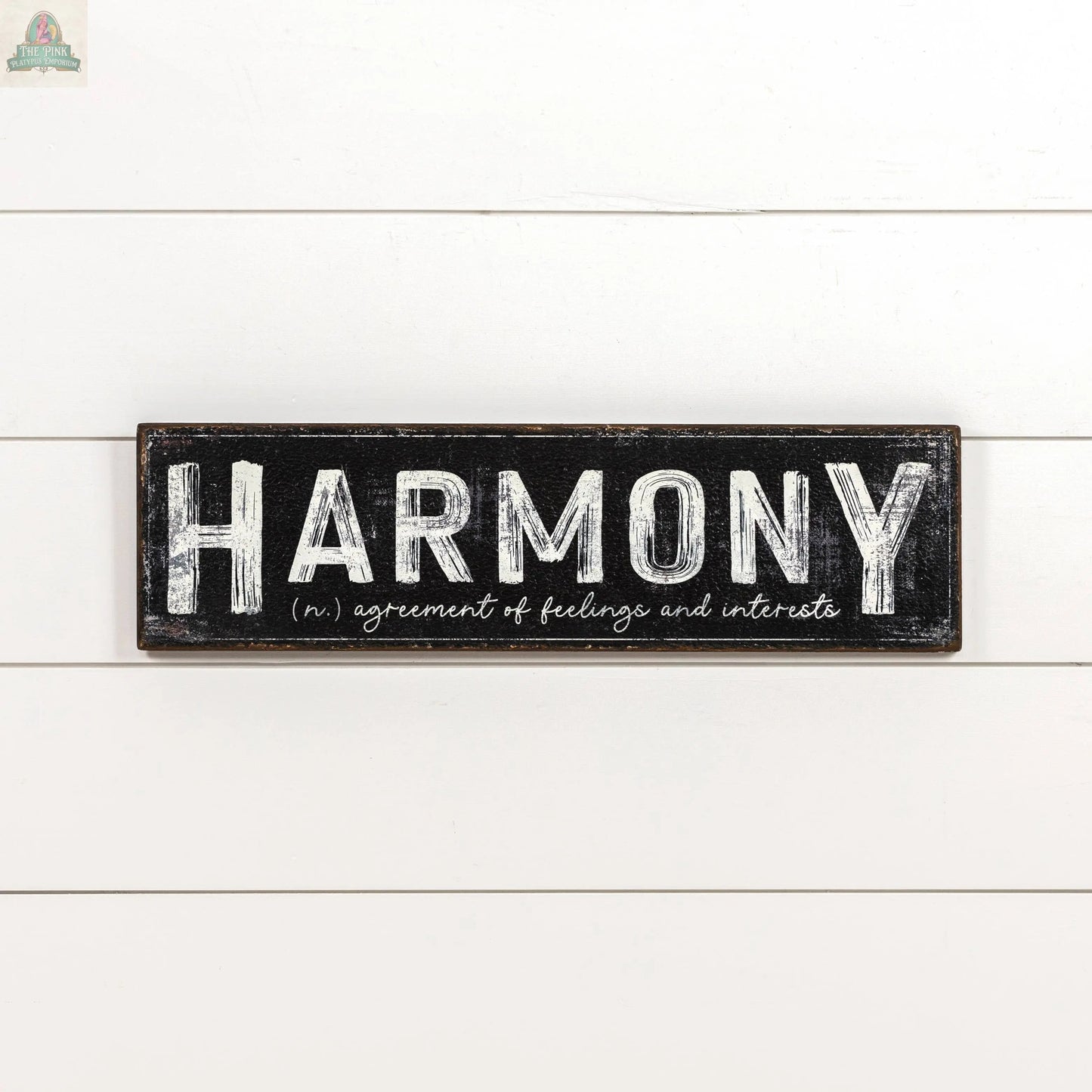 The 24" HARMONY METAL SIGN by Ragon House is a black rectangular metal sign with bold white "HARMONY" lettering and its meaning below, adding an elegant accent to your home decor.