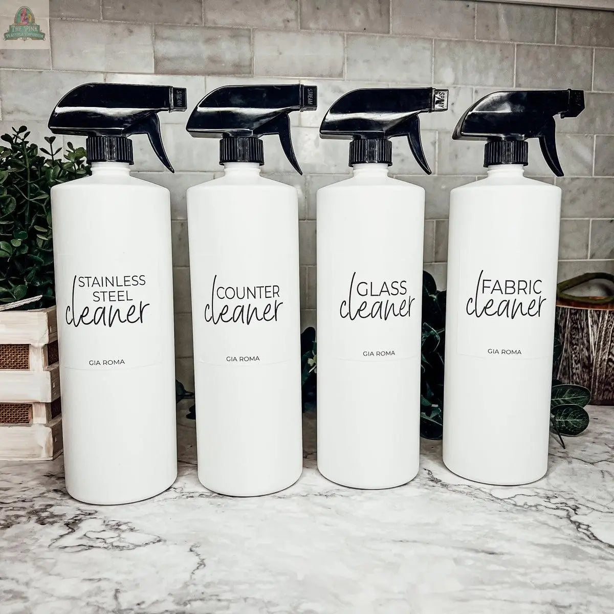Four 32oz Plastic Cleaning Bottles labeled "Stainless Steel Cleaner," "Counter Cleaner," "Glass Cleaner," and "Fabric Cleaner" stand in a row on a marble countertop, with waterproof labels, a refillable design, and green plants in the background.