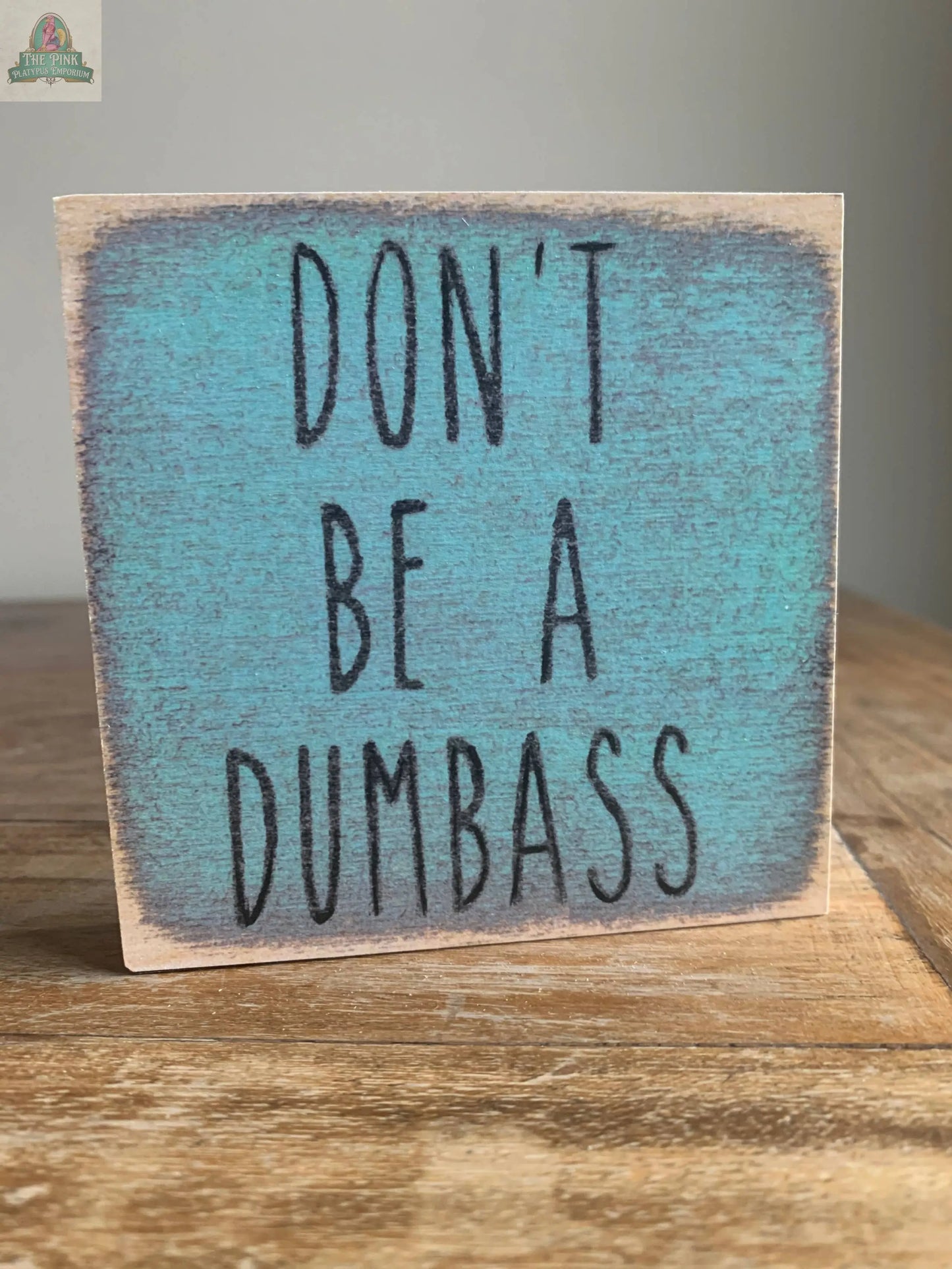 The 4X4-Dumbass block sign by Pink Platypus Emporium features “DONT BE A DUMBASS” in bold black letters on a distressed turquoise background, adding playful charm to your home décor.