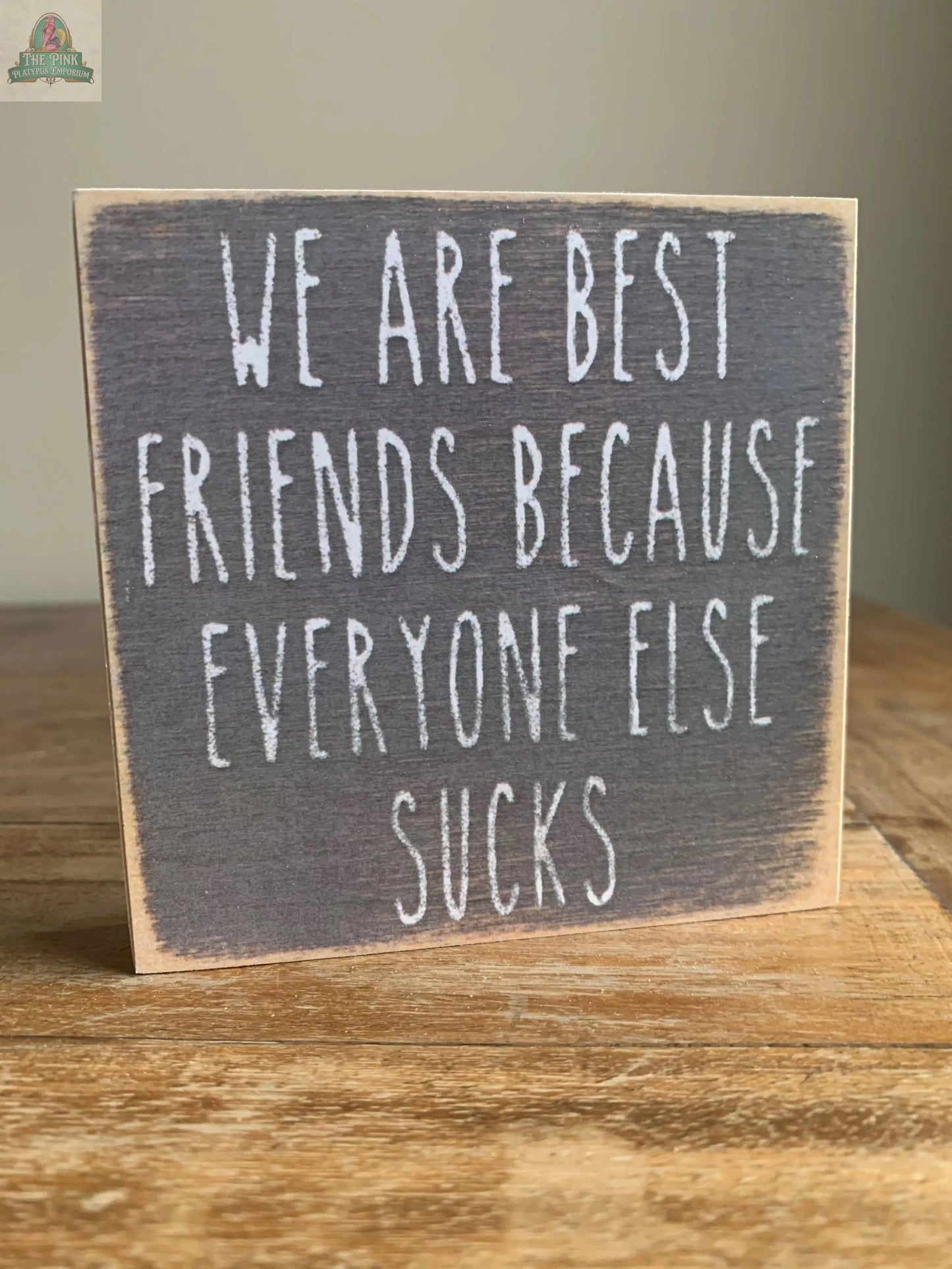 The Pink Platypus Emporium 4X4-Everyone Sucks wooden block sign features distressed edges and bold white text: "WE ARE BEST FRIENDS BECAUSE EVERYONE ELSE SUCKS." Ideal for adding a fun touch to your home decor.