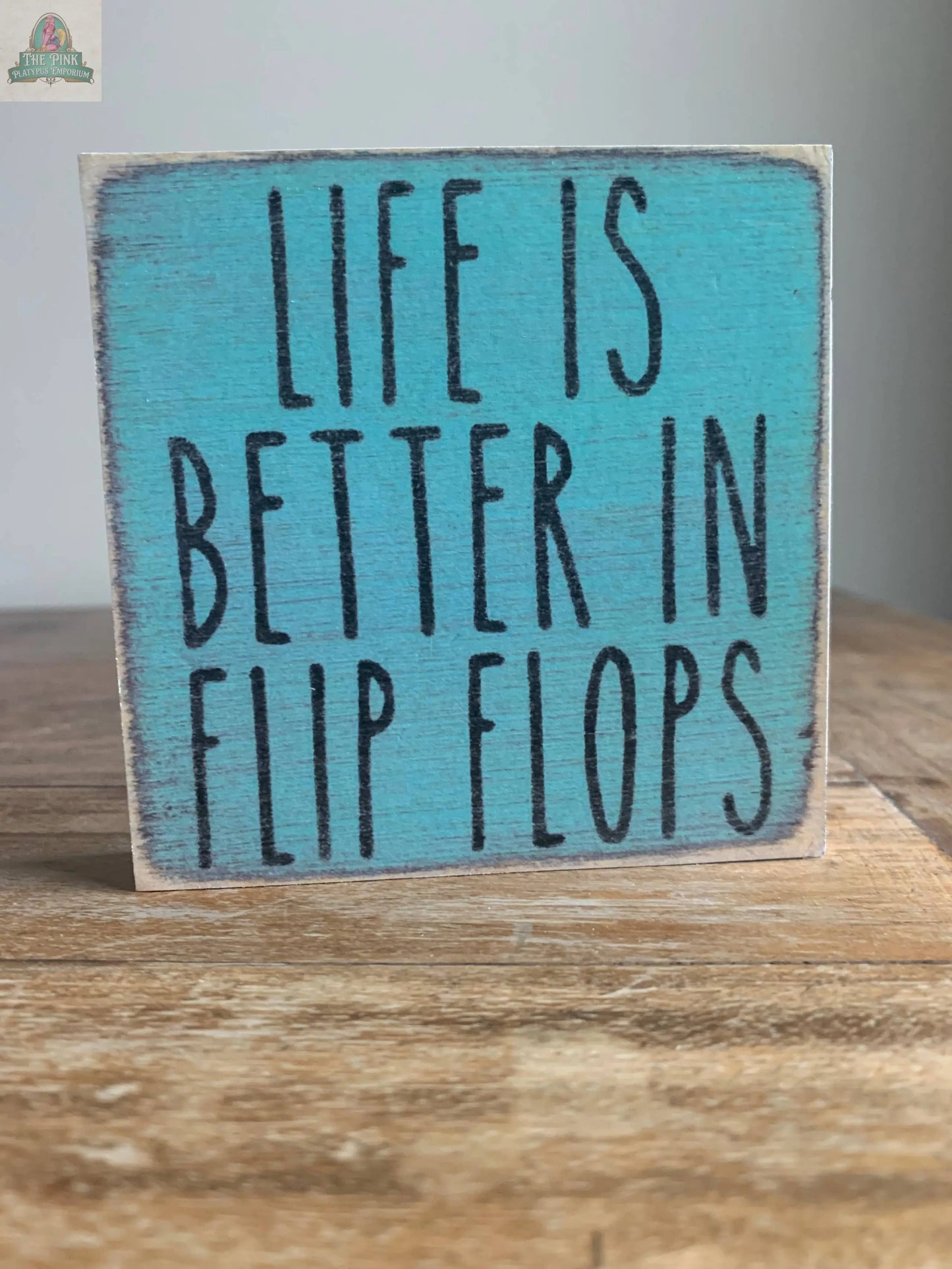 The Pink Platypus Emporium 4X4-Flip Flops wooden block sign features a distressed blue background and bold black lettering that says “LIFE IS BETTER IN FLIP FLOPS”—a charming home decor accent for any space.