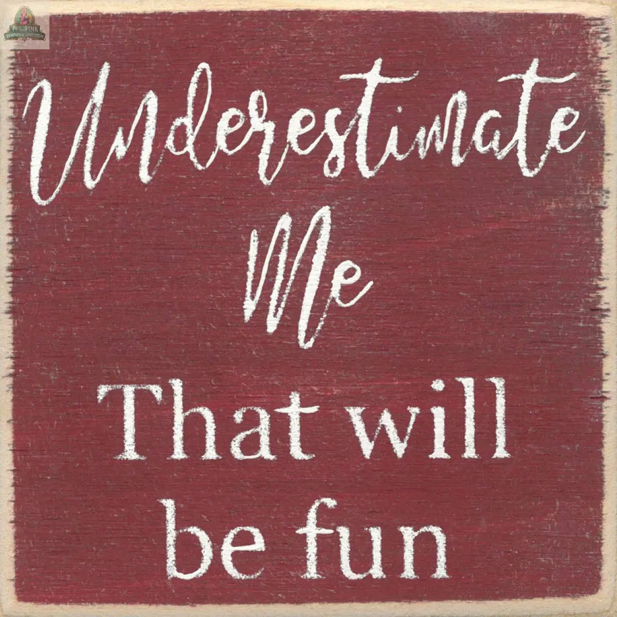 The 4X4-Underestimate Me sign from Pink Platypus Emporium features white cursive and block letters on a red, textured finish with uneven edges, giving it a rustic painted look. It reads: Underestimate me. That will be fun.