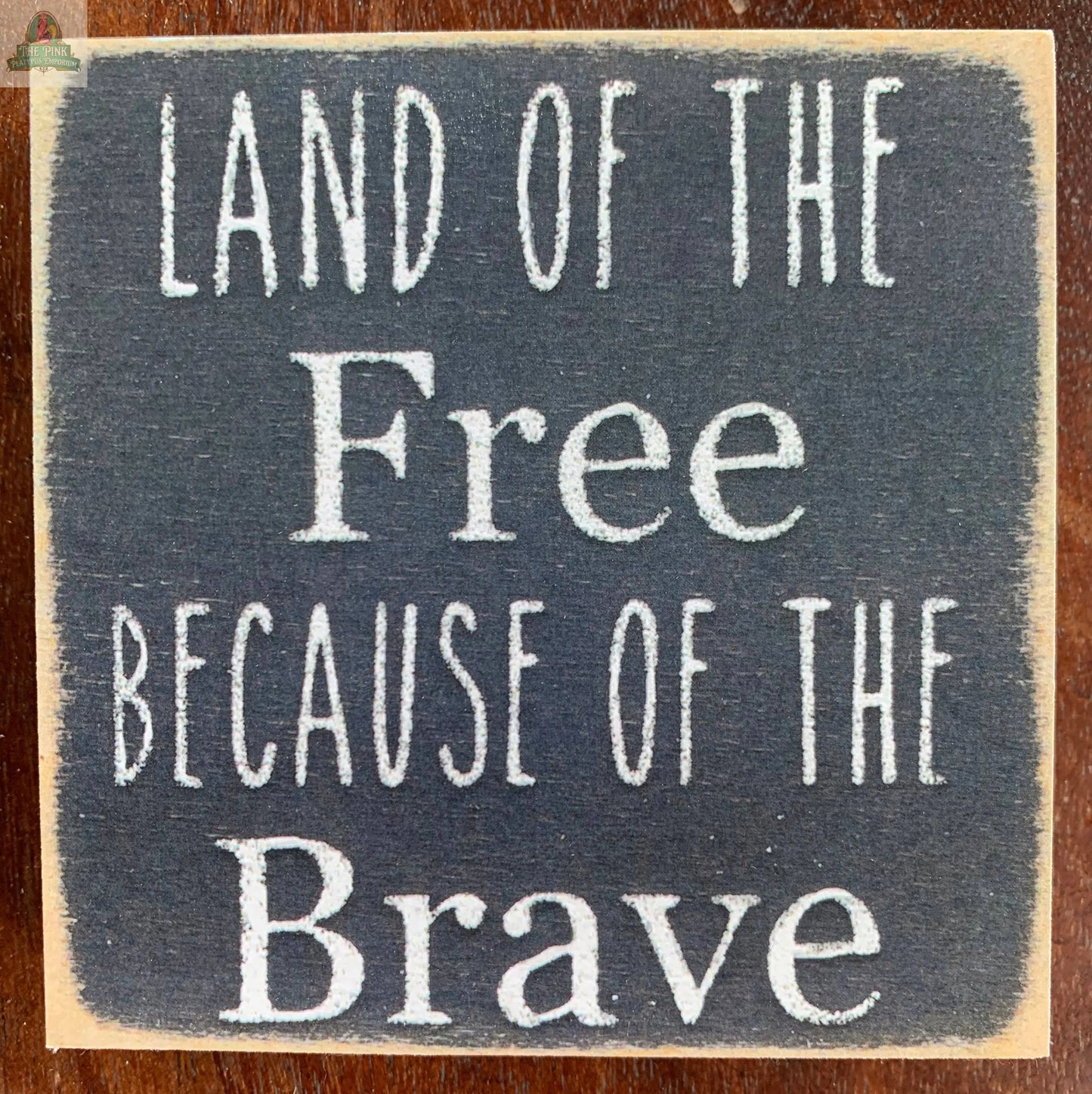 The Pink Platypus Emporium "Land of The Free Because of the brave" sign features white text on a dark background, adding handcrafted patriotic charm to any space.
