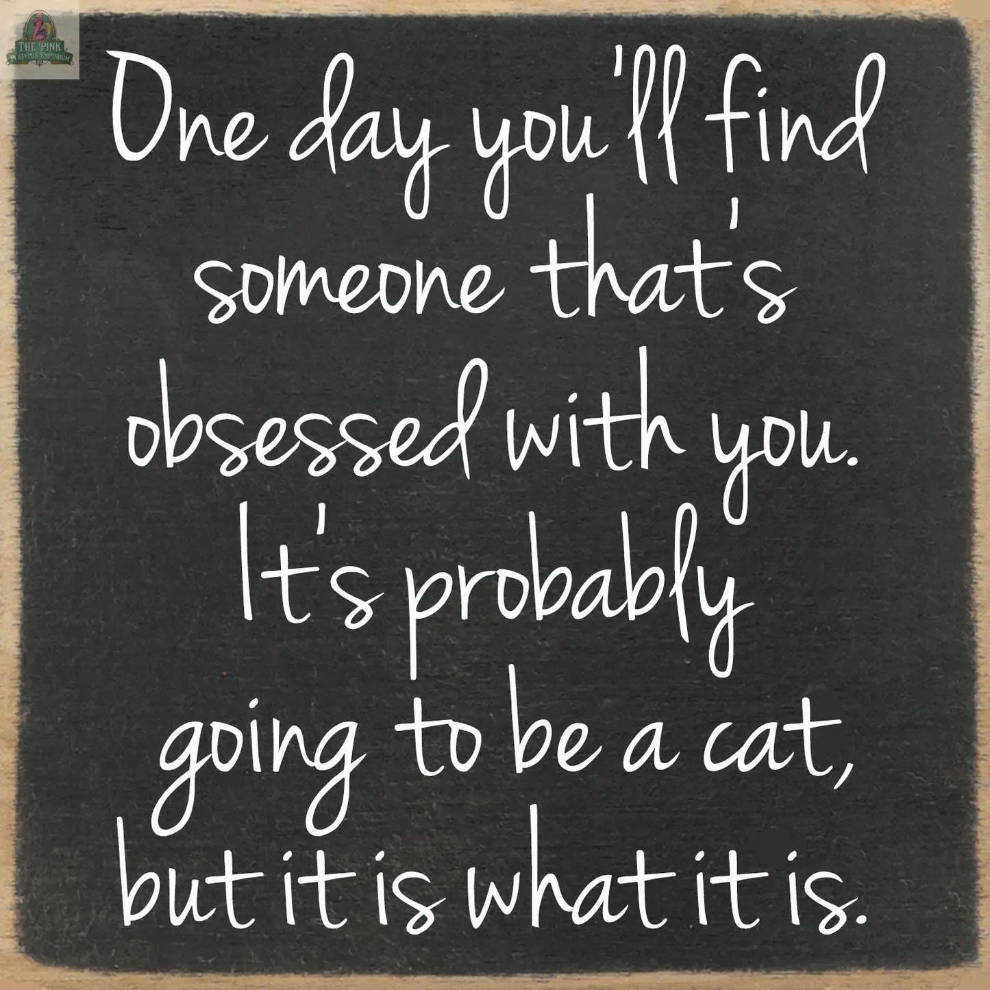 White handwritten text on a black chalkboard-style 5x5 block reads: One day you'll find someone obsessed with you. It's probably a cat, but it is what it is. A quirky sign by pink platypus emporium.