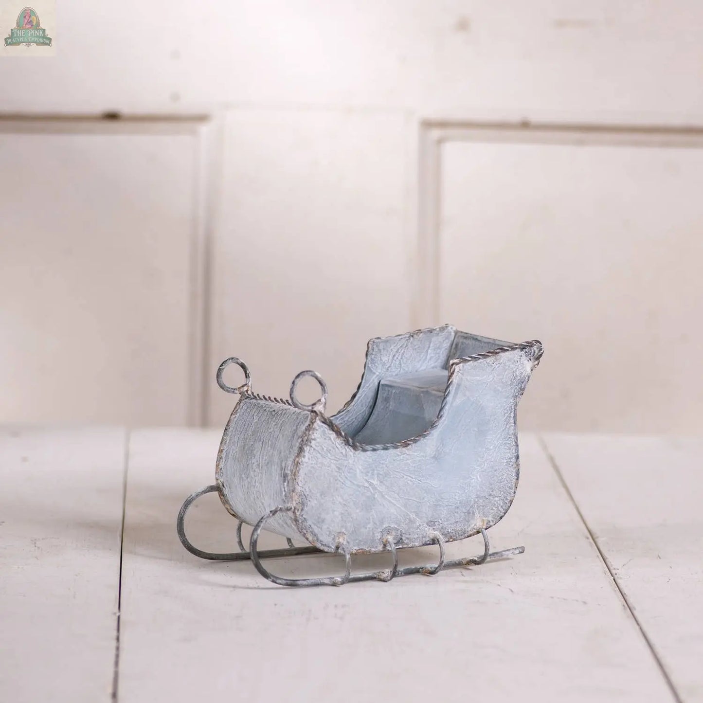 The Pink Platypus Emporium 7-Inch Tin Sleigh in Rustic White Wash adds classic holiday charm, featuring a vintage-inspired distressed finish. Perfect for decorating tables or mantels against light backgrounds.