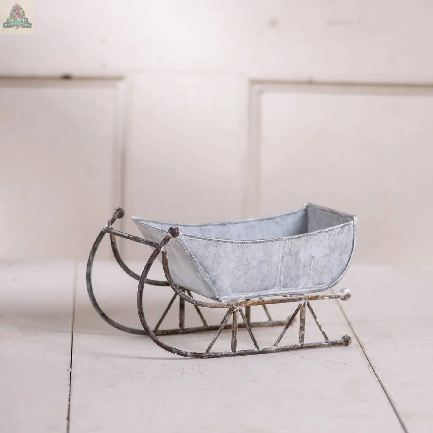 The Pink Platypus Emporium 9-Inch Tin Sleigh in Rustic White Wash, featuring curved runners and a weathered finish, sits on a white wooden surface—perfect for adding vintage charm to your rustic holiday decor.