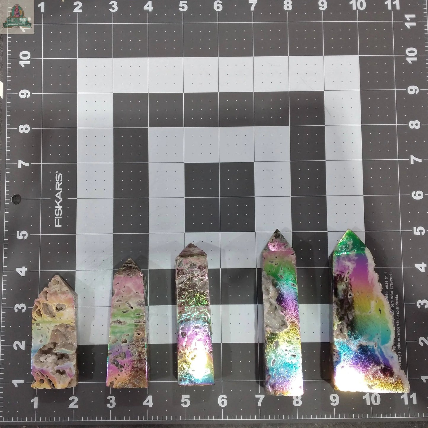 Five vibrant crystal points, including the Angel Aura Druzy Sphalerite Obelisk by Keystone Crystals, are lined up on an inch-marked gray grid mat. Each shimmering tower from Pink Platypus Emporium displays swirling pastel hues and metallic reflections.