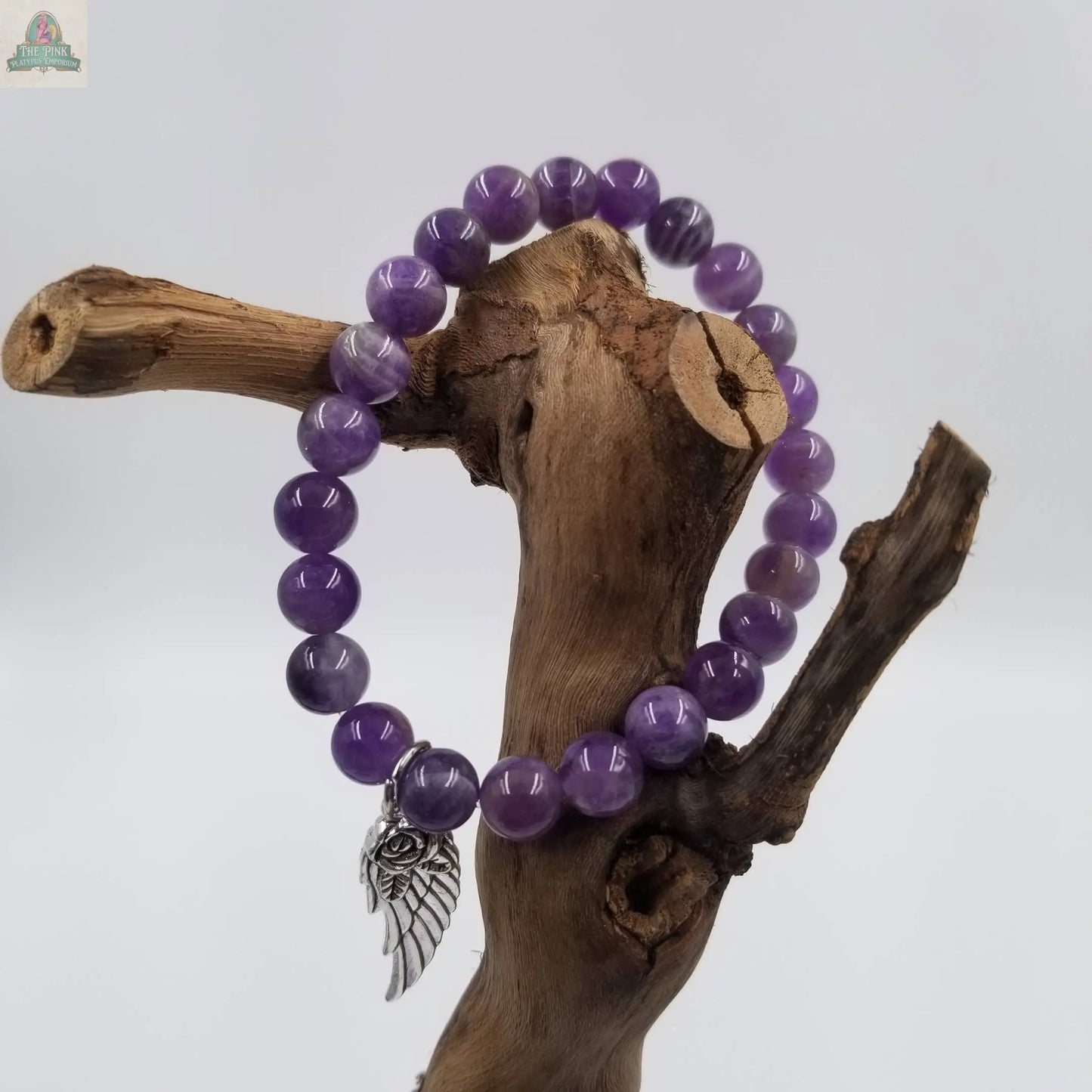 The Pink Platypus Emporium Angel Wing Amethyst Bracelet With Card, shown draped on a wooden branch, features soothing beads and a silver wing charmโan elegant Mother's Day gift and beautiful stress relief accessory.