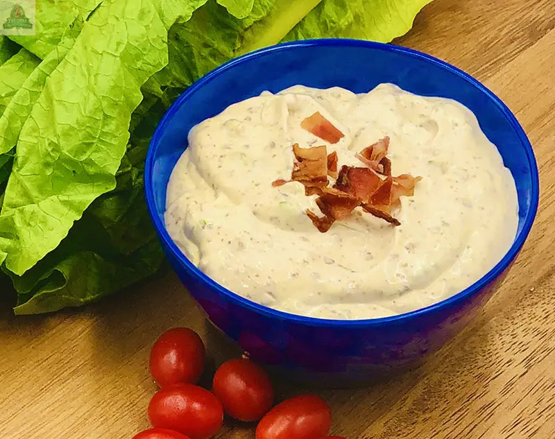 A blue bowl filled with Carmies Kitchen BLT Dip Mix, topped with chopped bacon, sits on a wooden surface surrounded by fresh romaine lettuce leaves and grape tomatoes.
