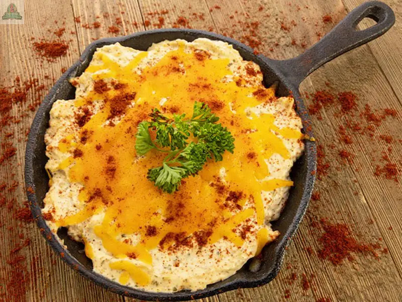 A small cast iron skillet of Carmies Kitchen Baked Enchilada Dip Mix, topped with shredded cheddar, paprika, and parsley, sits on a rustic wooden table sprinkled with paprika.