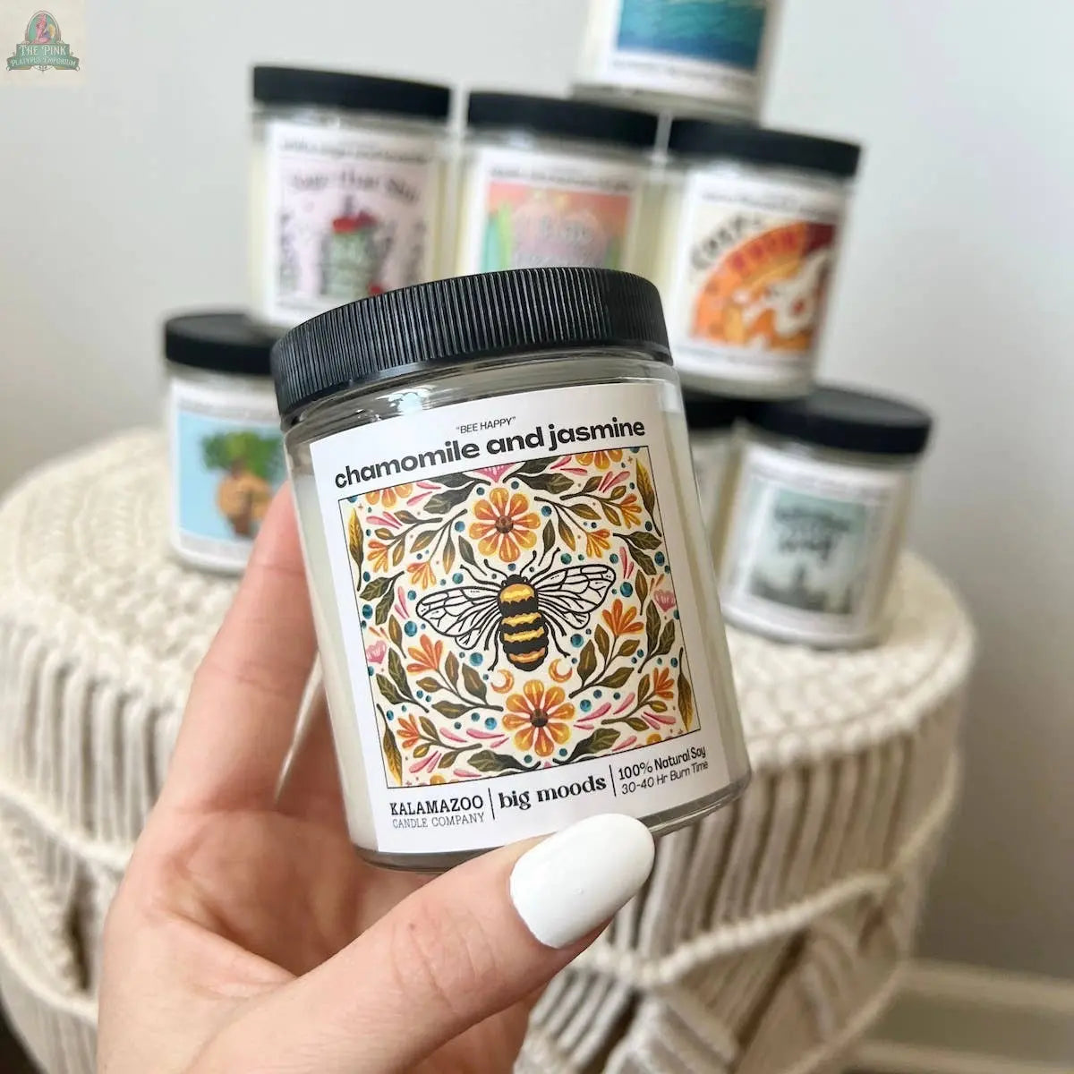 A hand with white-painted nails holds the Pink Platypus Emporium "Bee Happy" Chamomile and Jasmine Candle, made from natural soy wax and featuring a bee illustration; other colorful scented candles appear blurred in the background.