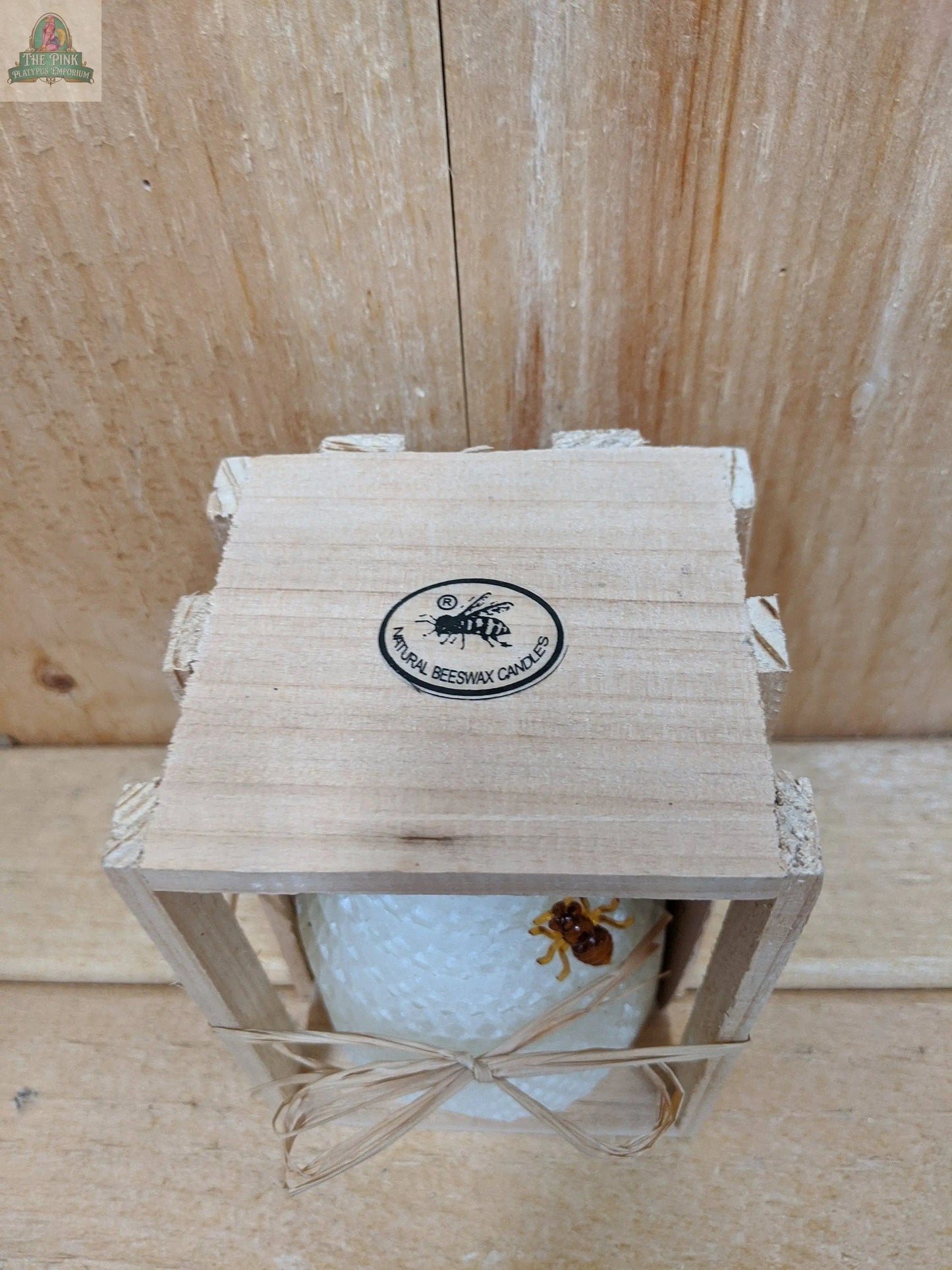 A handcrafted white Beeswax Honey Comb candle from Pink Platypus Emporium, featuring a honeycomb pattern and a decorative bee, sits in a small wooden crate. The background is wooden.