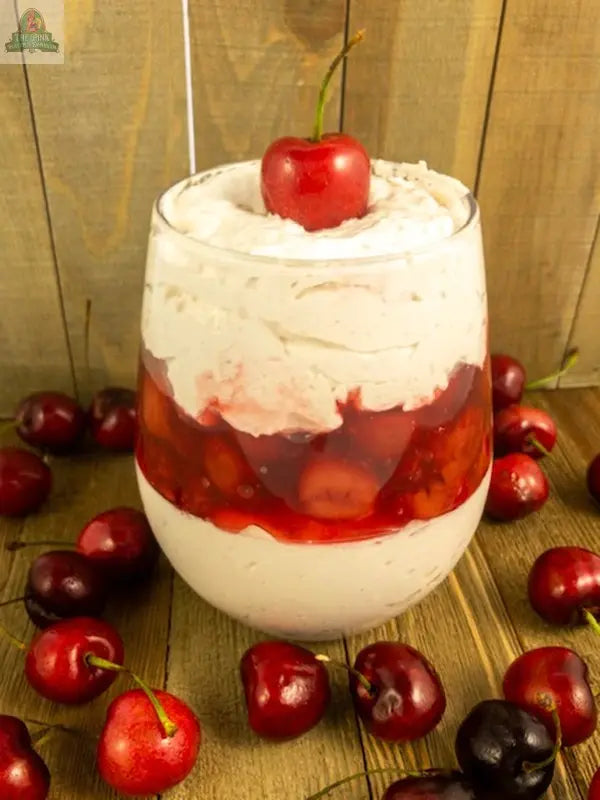 Carmie's Kitchen Black Cherry Vanilla Cheesecake Dip is a layered dessert in a glass with all-natural whipped cream and black cherry cheesecake filling, topped with a cherry and fresh cherries on a wooden surface.