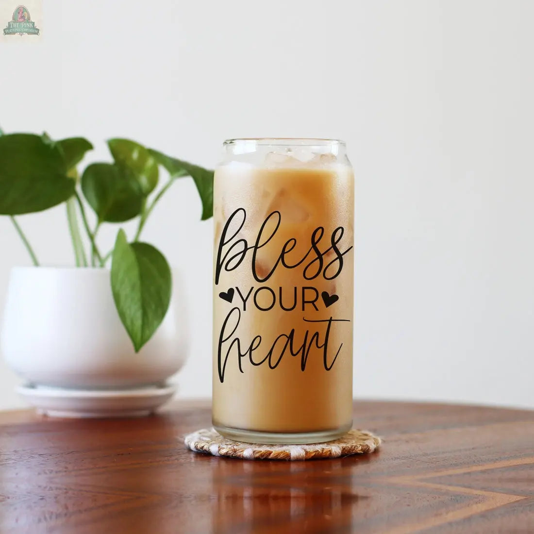 The Bless Your Heart 20oz glass cup with bamboo lid holds iced coffee on a coaster next to a white potted plant on a wooden table, set against a simple light background.
