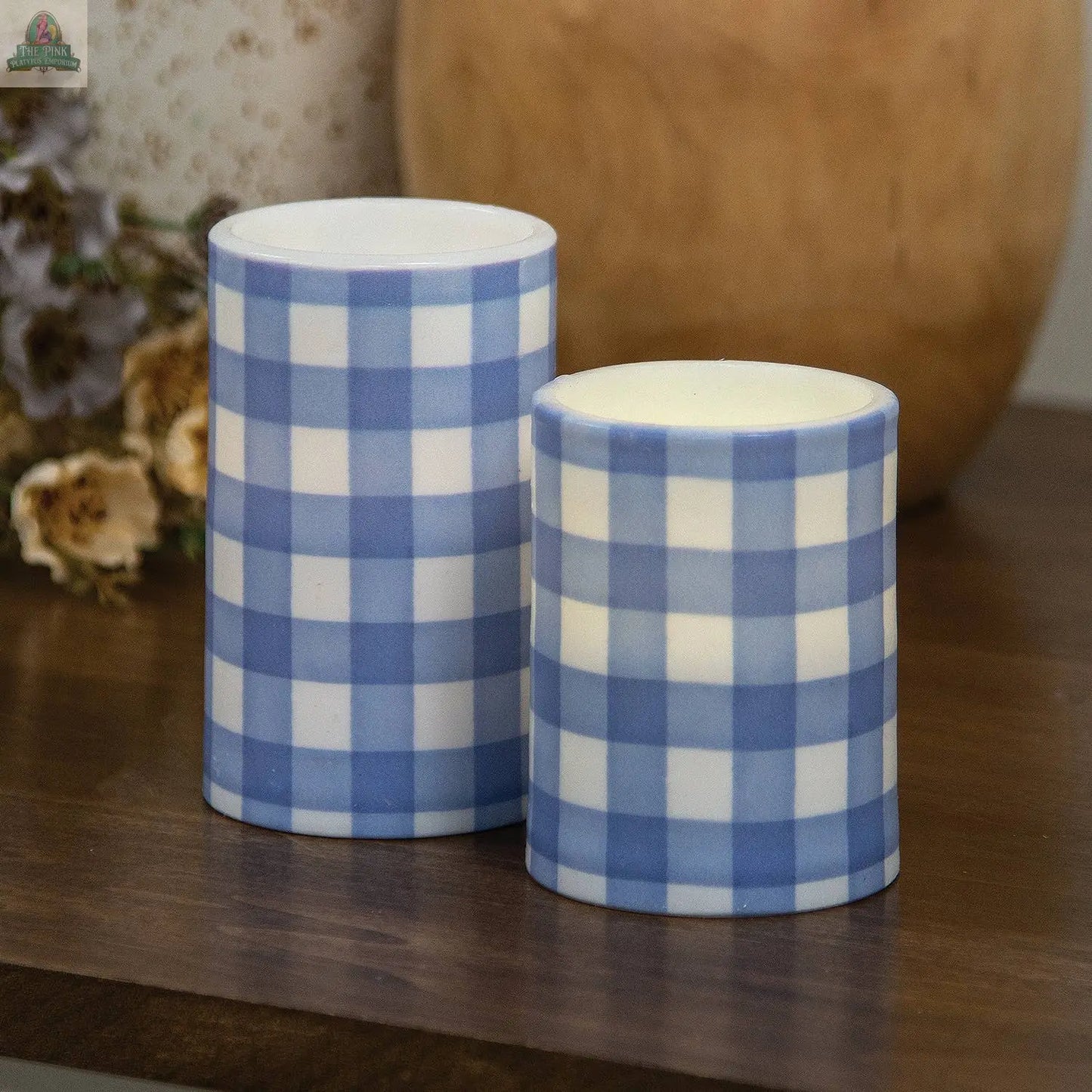 Two cylindrical ceramic containers with blue and white checkered patterns, inspired by Pink Platypus Emporium’s Blue Gingham Check Timer Pillar (3"x5"), sit on a wooden surface; one is taller, with dried flowers and a beige vase behind.