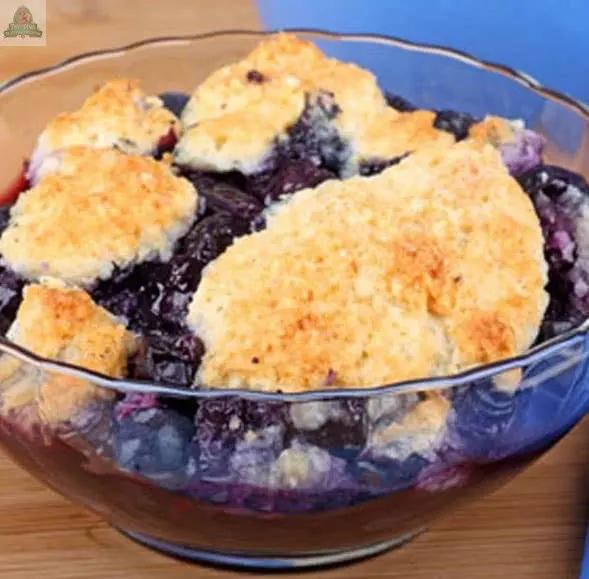 A clear glass bowl of blueberry cobbler with golden-brown biscuit topping sits on a wooden surface beside Blueberry Cobbler Atomizer Oils, filling the room with the comforting scent of freshly baked dessert.