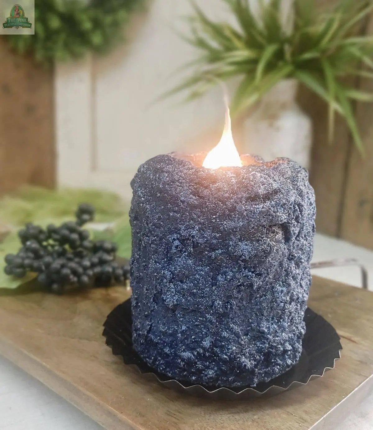 The Blueberry Cobbler Electric Candle, textured in dark blue, rests on a black tray atop wood, casting a cozy glow among greenery and faux grapes in the softly blurred background.