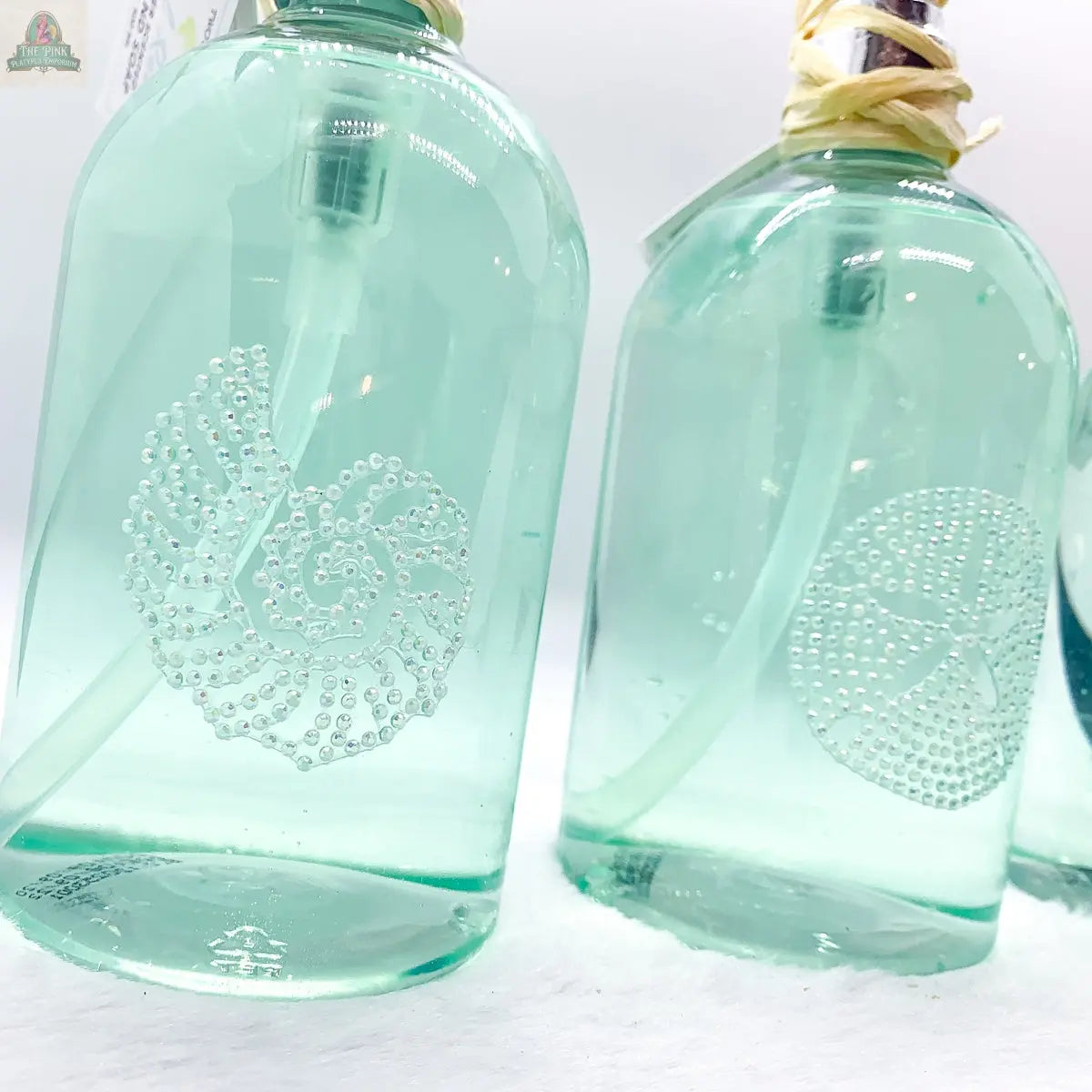 Two By the Sea Hand Soap dispensers, light green with embossed nautilus shell and sand dollar designs, sit on a white surface. Raffia-wrapped pumps make them perfect for beach-inspired bathroom decor.