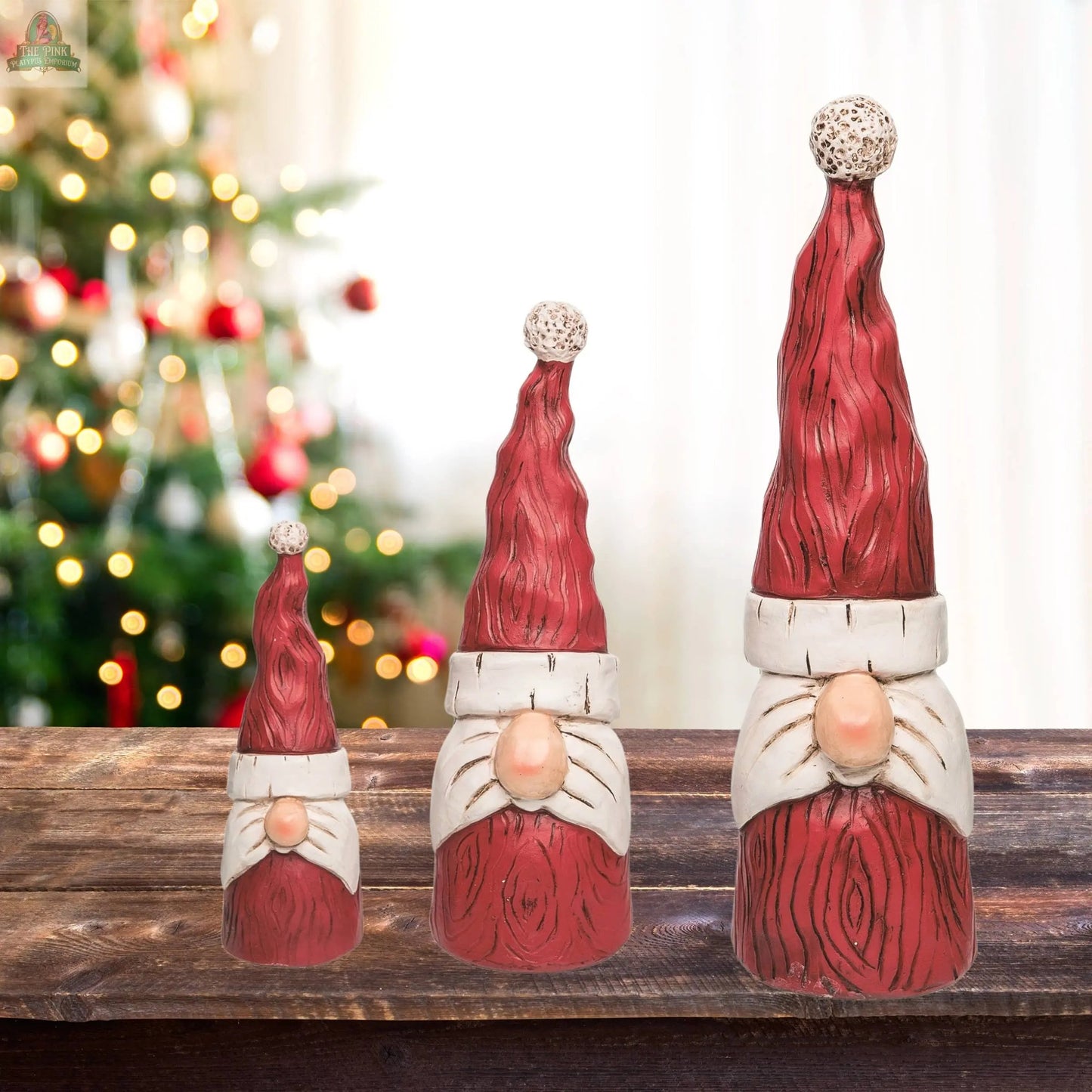 The CLOSEOUT Christmas Resin Gnome Santa Decor Set of 3 features gnome figurines with red hats and white beards, perfect for whimsical holiday décor. Display them on a tabletop with a festive, twinkling Christmas tree in the background.
