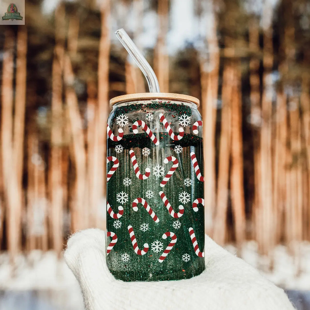 A gloved hand holds the Candy Cane holiday glass cup with lid and straw, featuring candy cane and snowflake designs, outdoors with tall, blurred pine trees in the background.