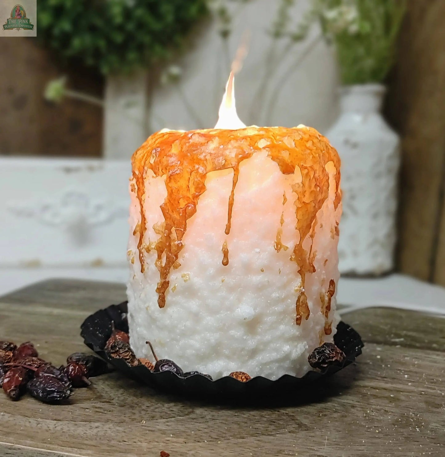 The Caramel Corn Electric Candle, handmade in the USA, displays white wax with melted amber drips on a black dish surrounded by dried berries atop wood, with a softly blurred white vase and greenery in the background.