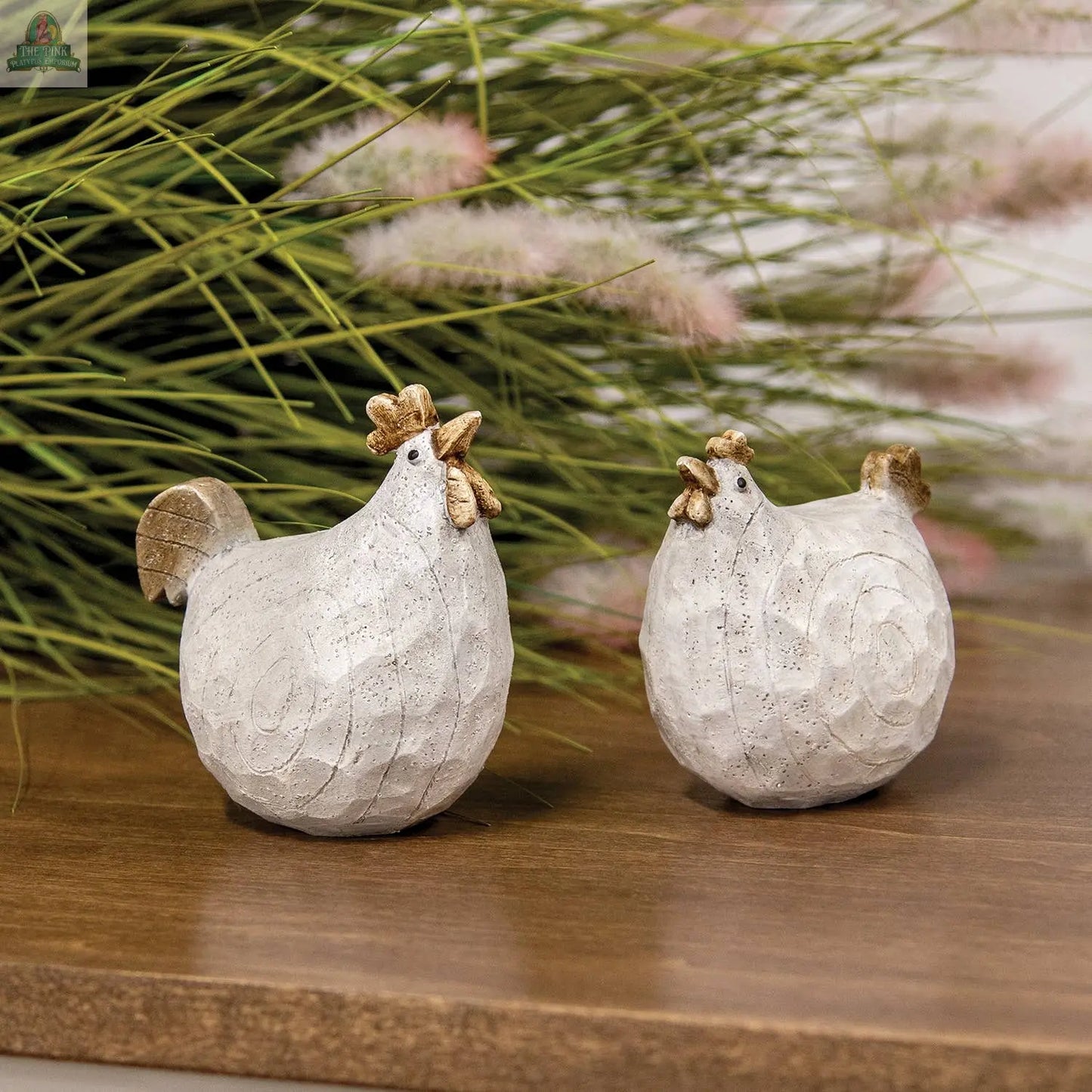 The Pink Platypus Emporium Carved Look Resin Chicken Sitter features two white chicken figurines with gold accents, displayed on wood before green grass—ideal farmhouse decor.