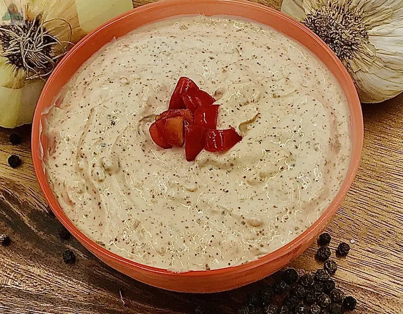 A bowl of Carmie's Kitchen Chipotle Ranch Dip Mix, garnished with chopped red peppers and surrounded by black peppercorns, onions, and garlic on a wooden surface.