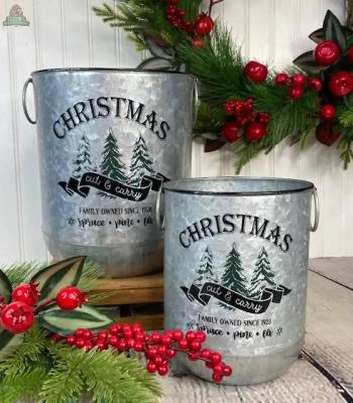 The Pink Platypus Emporium Christmas Cut & Carry Metal Bucket w/Ring Handles (Set of 2) features festive pine and red berry decor, creating a charming holiday centerpiece, with a garland in the background for added seasonal cheer.