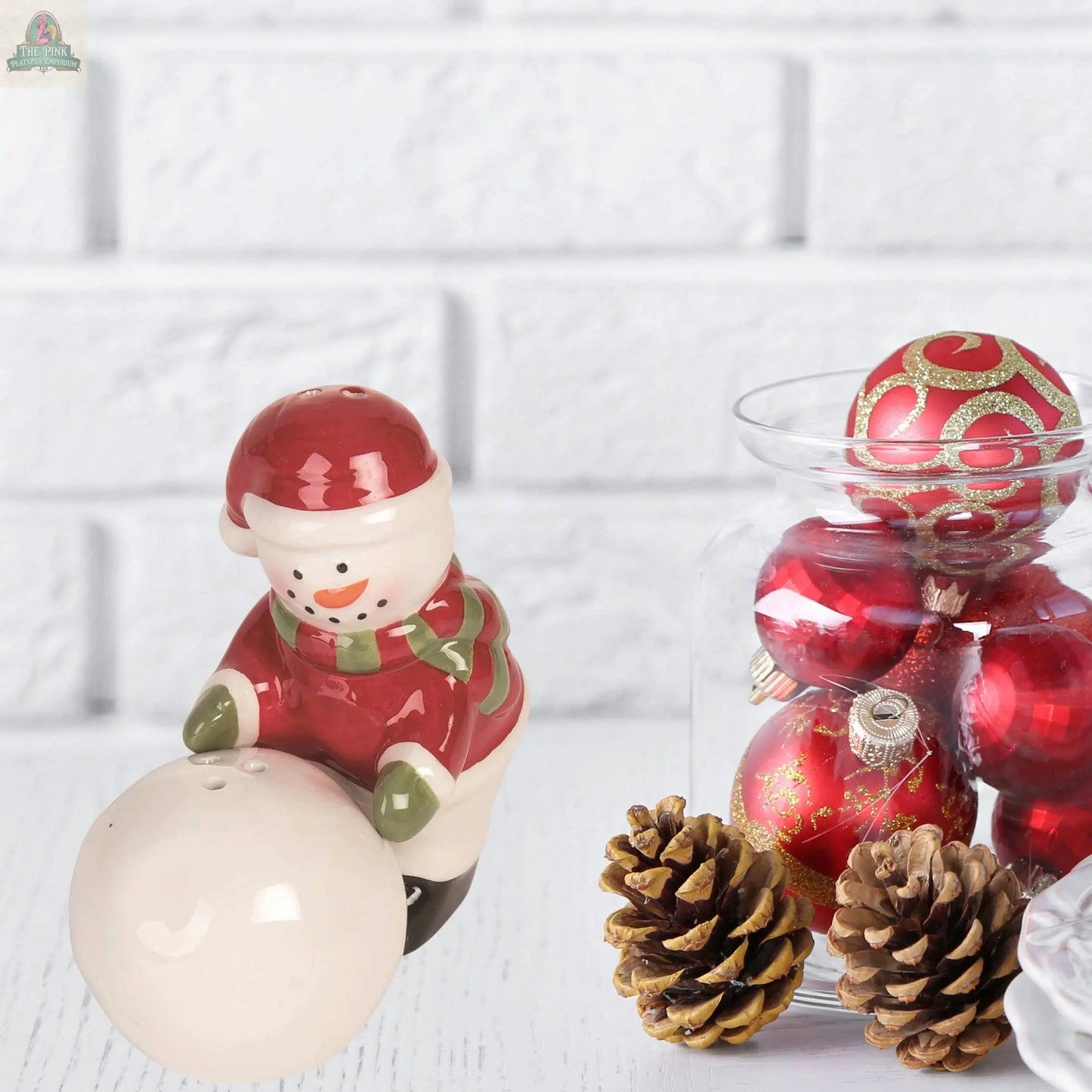 The Christmas Snowman Snowball Salt Pepper Shaker Set (2 pcs, ceramic) in festive red and green sits beside a jar of red ornaments and pinecones against a white brick background—an ideal, charming holiday gift.