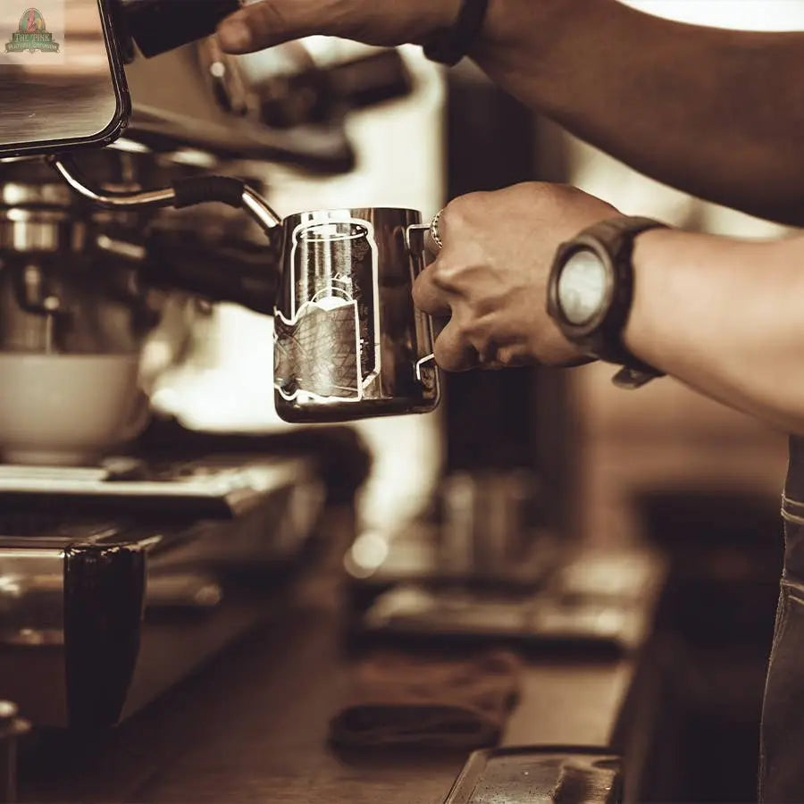 A barista prepares coffee at an espresso machine, holding a metal milk frothing pitcher. Warm tones evoke the cozy scent of the Coffee House Hearth Candle by Pink Platypus Emporium. The barista is wearing a watch.