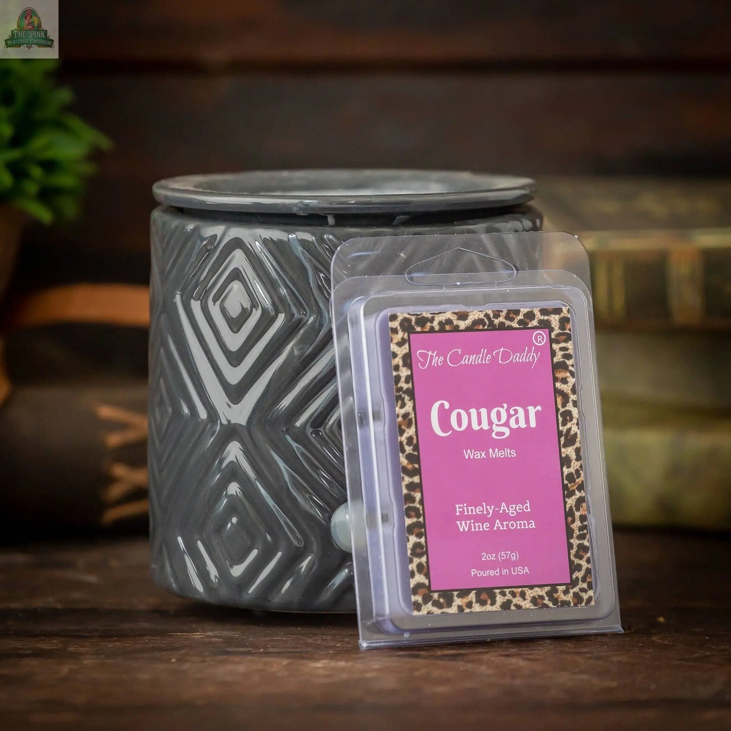 A gray patterned wax warmer sits beside a pack of Cougar - Fine Wine Scented Wax Melts Cubes (2oz, 1 Pack) in purple and leopard-print packaging on a wooden surface, with blurred books and greenery in the background.
