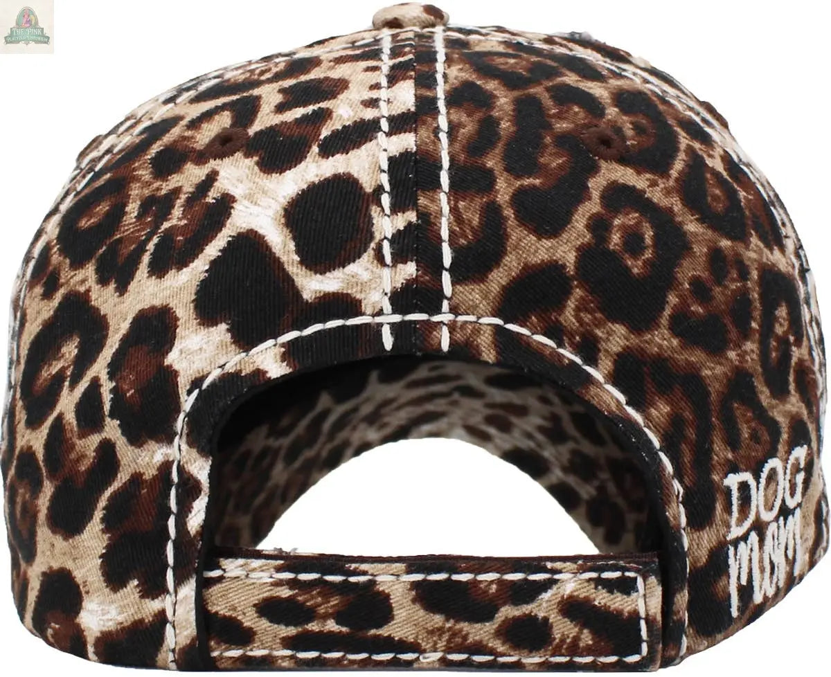 A close-up shows the back of the Pink Platypus Emporium DOG MOM WASHED VINTAGE BALLCAP: MDM, featuring a brown and black leopard print, visible white stitching, and partial white “DOG” embroidery on the right side.