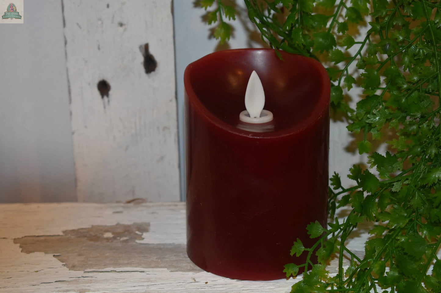 The Pink Platypus Emporium Moving Flame LED Candle Deep Red 3x4in is displayed on a weathered white wooden surface beside a green leafy plant, set against a distressed white wooden background.