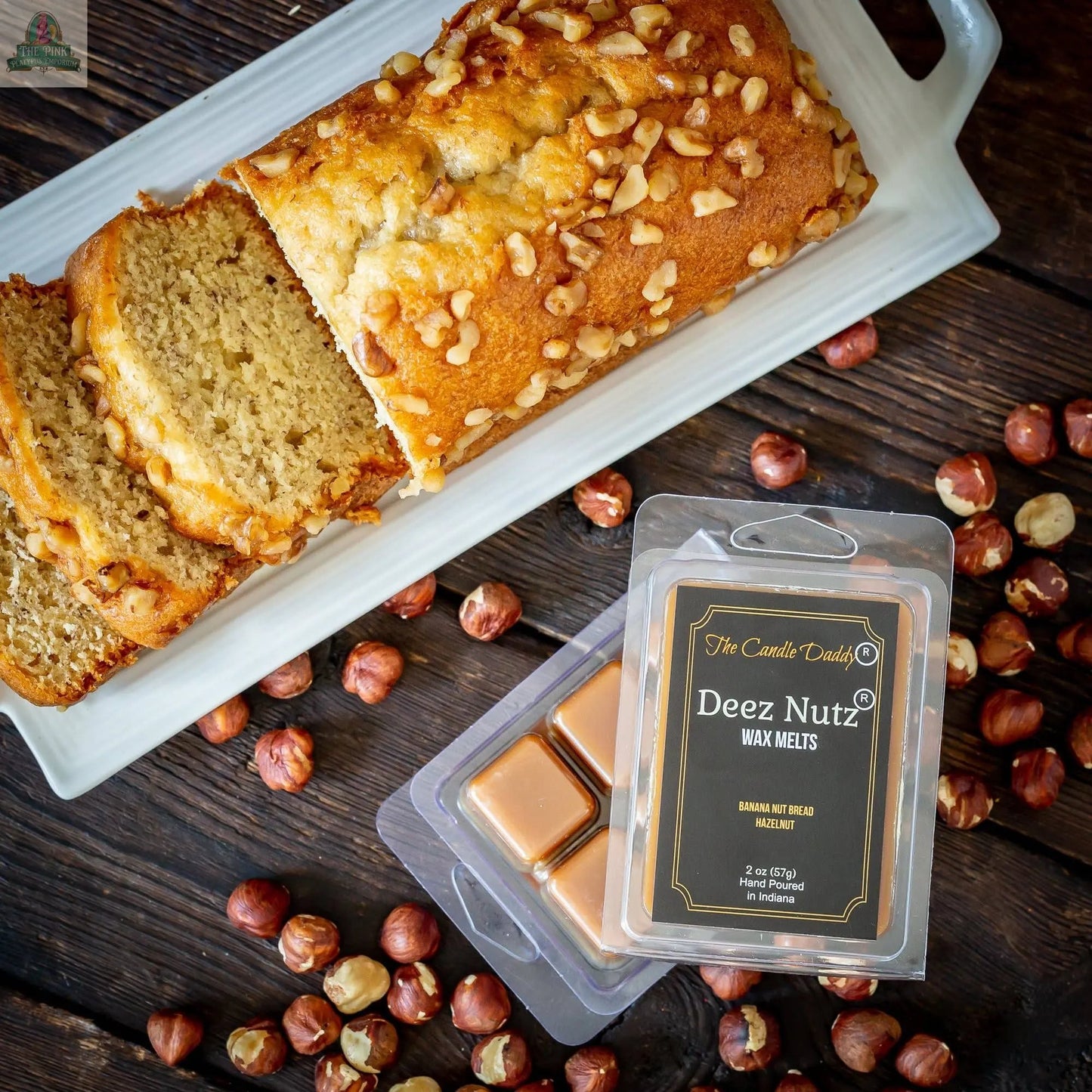 A partially sliced nut bread sits on a white plate with hazelnuts, next to a pack of Deez Nutz Wax Melts - Banana Nut Bread, featuring tan, scented wax cubes.