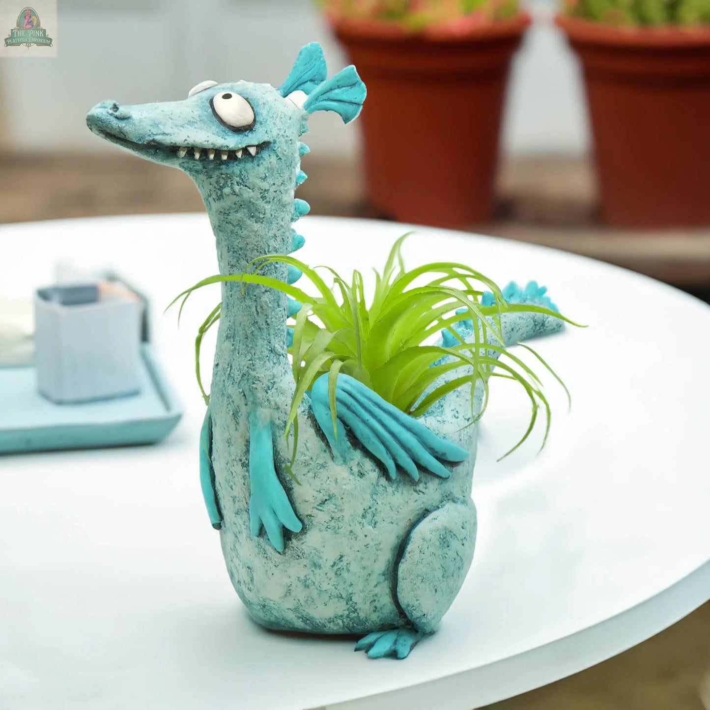 The Drake Teal Dragon Blobhouse Planter by Blob House Planters holds a small green plant on a white table with blurred pots behind—ideal for adding playful dragon planter charm to any whimsical home décor.