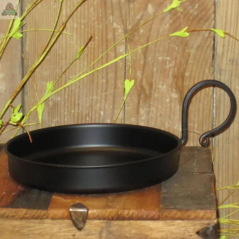 The Edged Black Metal Candle Pan w/ Handle rests on a rustic wooden surface, surrounded by green leafy branches and set against a wooden plank backdrop.