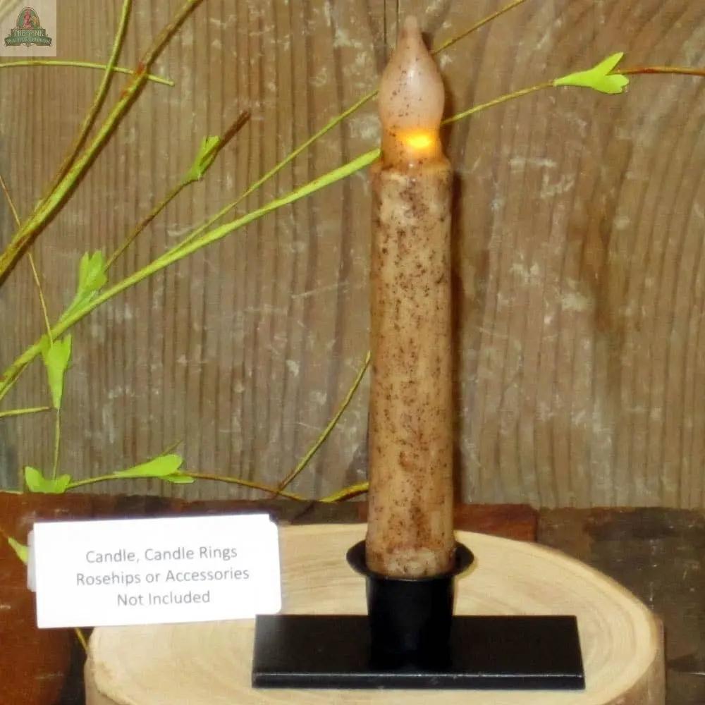 A tall, brown candle glows in a Flat Base Taper Candle Holder on a wooden surface. A sign notes: Candle, Candle Rings, Rosehips or Accessories Not Included. Green stems decorate the background.