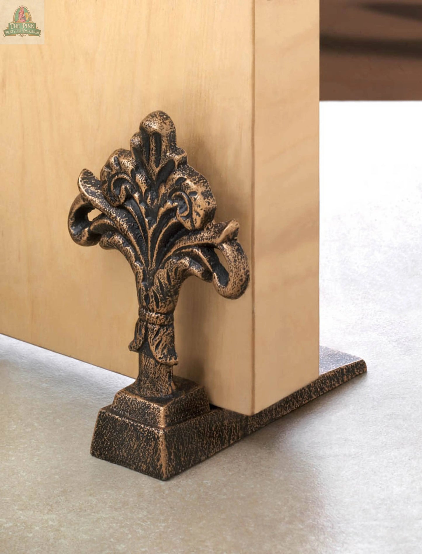 The Fleur De Lis Door Stopper, featuring a decorative antique-style floral design in cast iron, securely holds open a light wood door on a smooth, light-colored floor.