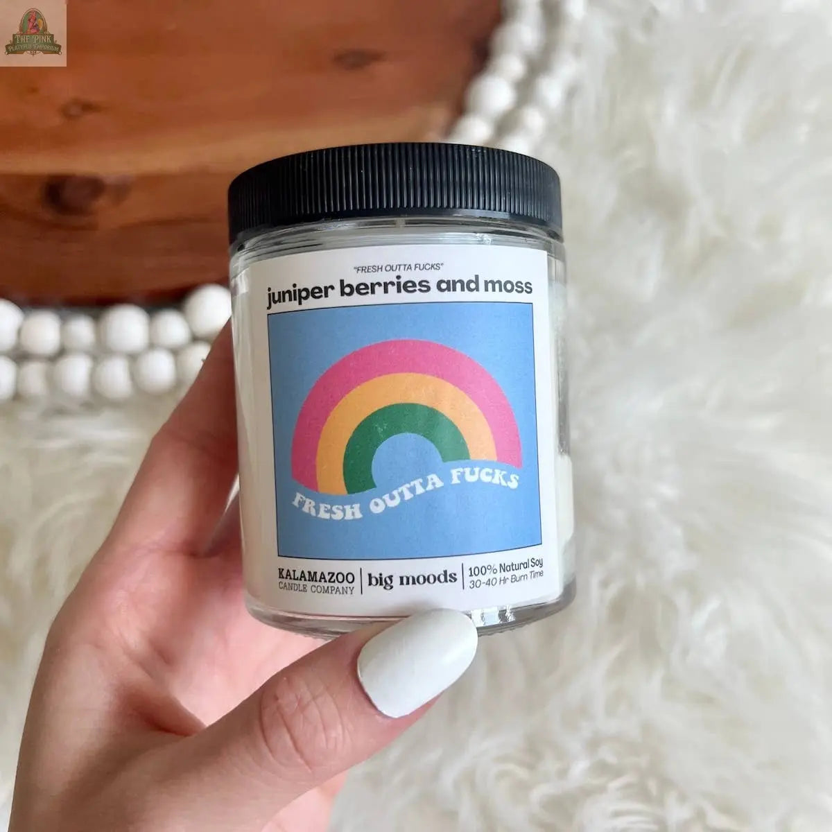 A hand with white-painted nails holds Pink Platypus Emporium's "Fresh Outta Fucks" Juniper Berries & Moss Soy Candle, featuring a rainbow label. The candle is on a wooden surface with white beads—perfect for natural aromatherapy.
