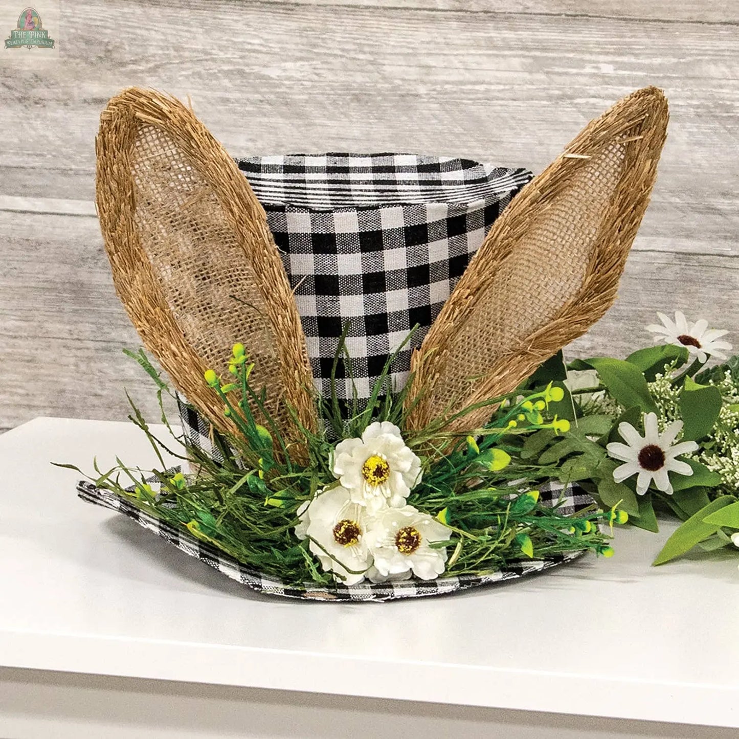 The Gingham Bunny Fabric Floral Top Hat by Pink Platypus Emporium, featuring woven bunny ears, white faux flowers, and green foliage, brings festive charm to your Easter decor as a delightful piece of spring floral décor.