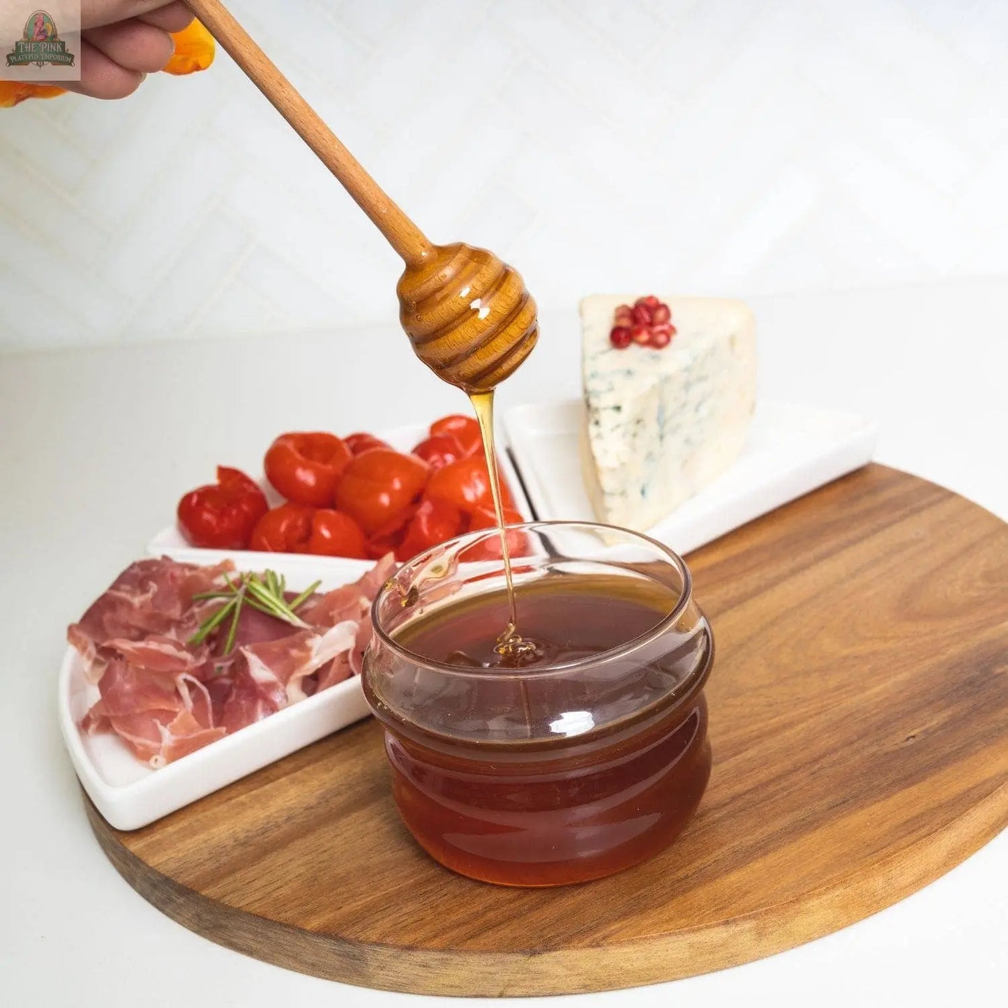 A hand drizzles honey from the Pink Platypus Emporium Glass Honey Jar & Dipper With Birch Wood Lid, while plates of cherry tomatoes, prosciutto, and blue cheese with pomegranate seeds rest on a round wooden charcuterie board.