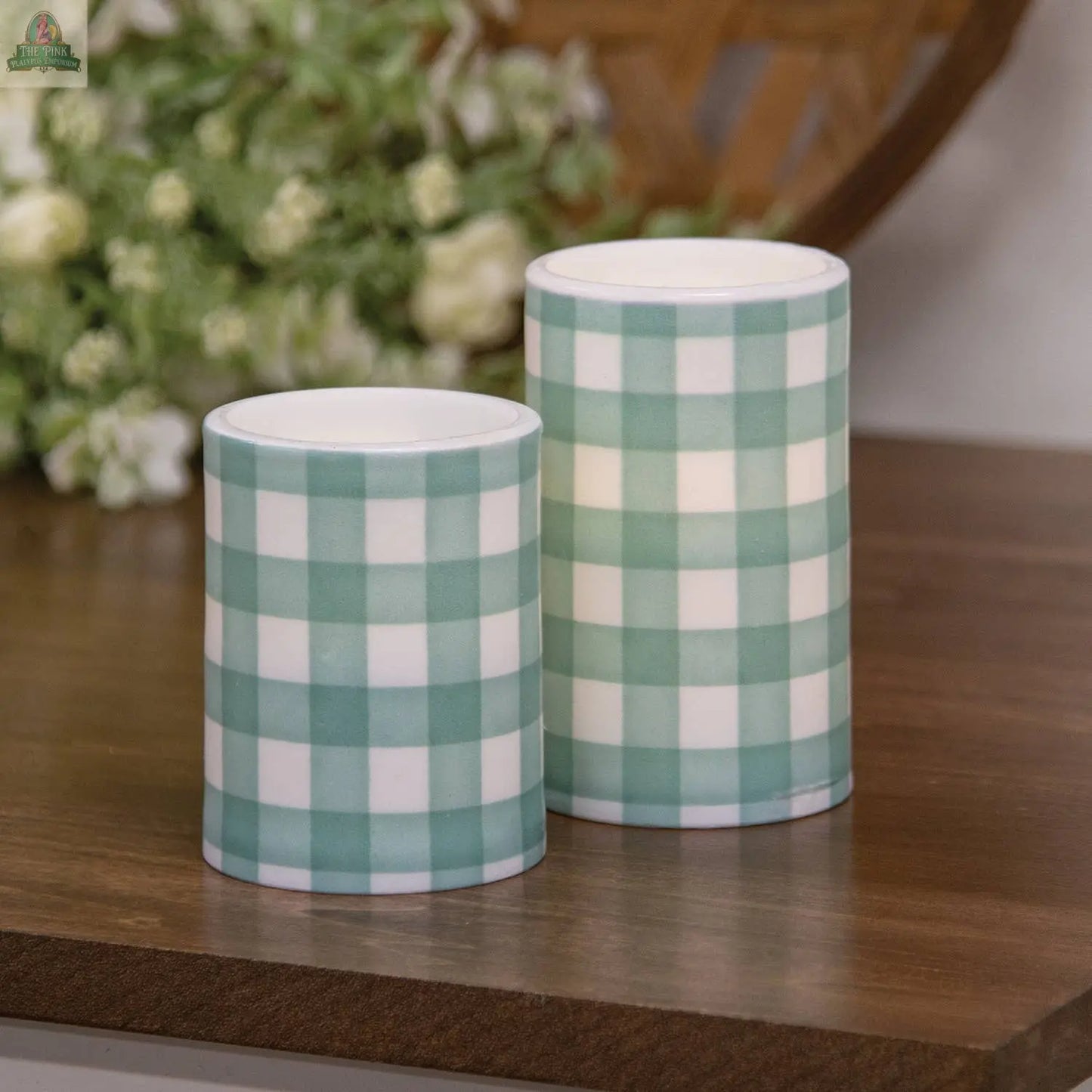 Two Pink Platypus Emporium Green Gingham Check Timer Pillar candles (3"x5") sit on a wooden surface, perfect for spring decor. Their green and white check pattern shines with blurred flowers and greenery in the background.