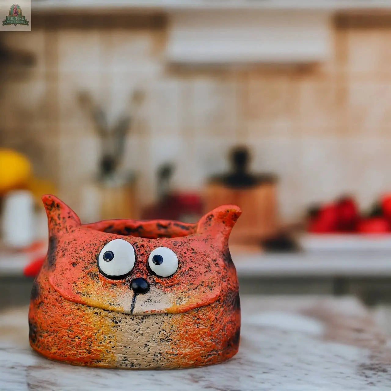 The Harley Tiger Cat Blobhouse Planter by Pink Platypus Emporium, shaped like a quirky orange feline with big googly eyes, sits on a marble countertop in a softly blurred kitchen setting.
