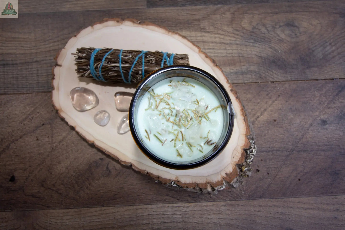 A small glass jar of Healing lotion, creamy and topped with dried herbs, rests on a wooden slab beside three clear stones, a citrine intention candle, and a blue-tied bundle of dried twigs on a wooden surface.