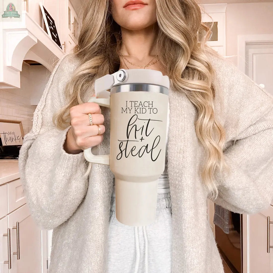 A woman with long, wavy blonde hair holds a large 40oz Hit & Steal baseball mom tumbler in her cozy kitchen. Dressed in a beige sweater with rings, she radiates sporty, laid-back mom vibes.
